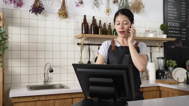 happy Asian coffee shop owner is using cash register while talking on the mobile phone with customers to take preorders for them at daytime.