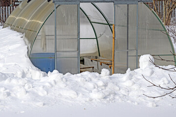 The greenhouse on the garden plot is covered with snow in early spring