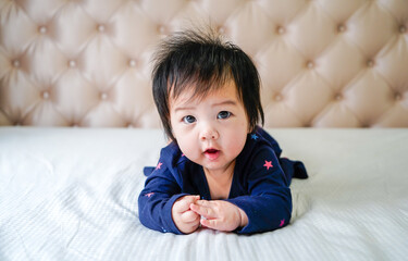 Cute little baby girl, playing at home in bed. Cute little caucasian baby lying on bed at home. Little cute baby girl, child in blue dress, smiling happily at camera in white sunny, bright bedroom.