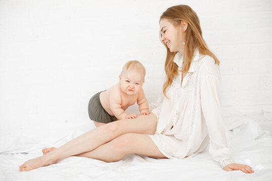 Young barefoot smiling woman in shirt with naked baby in nappy sitting white bedroom. Child development, mom relations