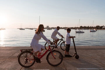 Fototapeta premium Happy family enjoying a beautiful morning by the sea together, parents riding a bike and their son riding an electric scooter. Selective focus 