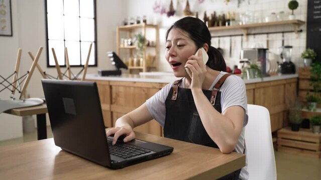 cheerful asian woman restaurant manager accepting preorder on mobile phone call and writes it down on computer in a closed store interior during the pandemic