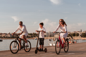 Happy family enjoying a beautiful morning by the sea together, parents riding a bike and their son riding an electric scooter. Selective focus 