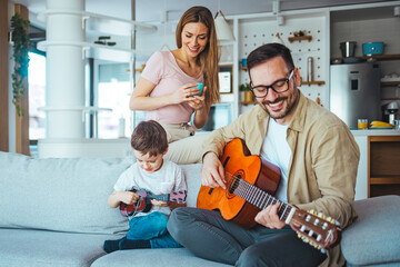 Father teaching his son to play on guitar at home. Son play on ukulele - hawaiian guitar. Father is...