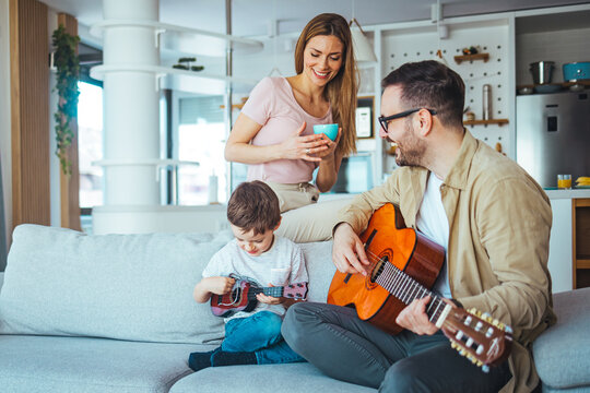 Shot of a father teaching his son the guitar. Father teaching his little son to play guitar at home. Man is istting on sofa with his son. He holds guitar. Man is pointing on strings. Boy looks at it