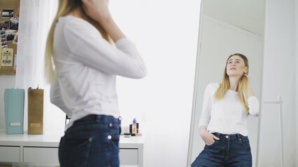 A young woman looks in the mirror, preens herself, admires herself, corrects her clothes and hairstyle. He smiles and likes himself.