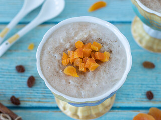 Homemade white rice pudding in a ceramic bowl topped with ground cinnamon and chopped dried apricots on a blue wooden table with raisins and spoons. Top view, a closeup. 