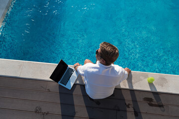 A modern elderly man enjoys the pool while working on his laptop next to a modern luxury house....
