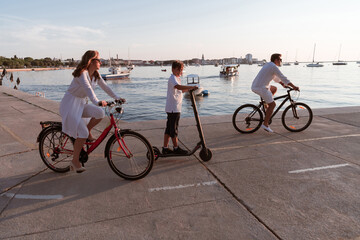 Fototapeta premium Happy family enjoying a beautiful morning by the sea together, parents riding a bike and their son riding an electric scooter. Selective focus 