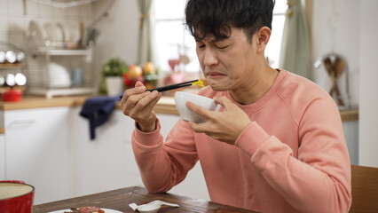 portrait of asian father making expression of disgust while tasting food with chopsticks and bowl...