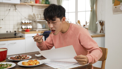 portrait of busy asian work from home father reading business documents while eating lunch at same...