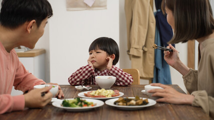 picky Asian boy shaking head while refusing food his father and mother put in his bowl during lunch time in dining room at home - obrazy, fototapety, plakaty