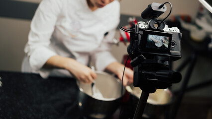A young woman blogger is interferes cream and recording videos for a food channel. A female pastry chef vlogs with her video camera mounted on a tripod in the kitchen.
