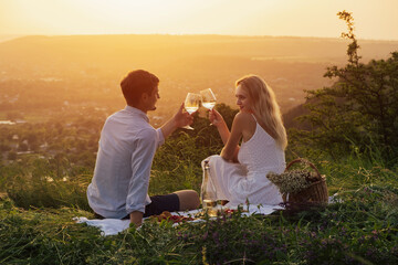 Couple in love on a white plaid take a picnic against the backdrop of a sunset in the mountains. Romantic time. Beautiful couple clinking glasses with wine..