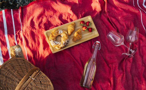 Romantic Picnic With Food And Drink On Red Blanket In The Sunny Summer Day. Top View. Croissants, Baguettes In Basket, Wine Bottle And Glasses. 