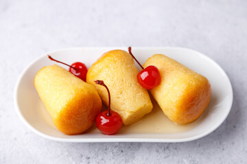Neapolitan Rum baba (or baba au rum) on a white plate with a cocktail cherry on a gray background. Small yeast cakes soaked in rum syrup. Traditional Italian pastry. Close-up, selective focus. - obrazy, fototapety, plakaty