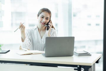 Cheerful young Asian woman smiling and looking away while having smartphone conversation in office