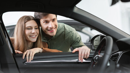 Cheerful young man and woman buying new car, checking automobile salon through window, panorama
