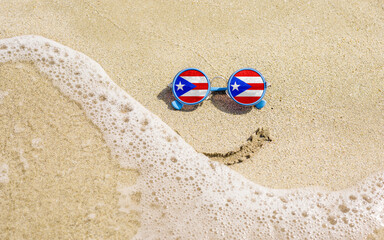 Fototapeta na wymiar Sunglasses with flag of Puerto Rico on a sandy beach. Nearby is a sea lightning and a painted smile. The concept of a successful vacation in the resorts of Puerto Rico.