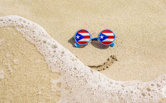 Sunglasses with flag of Puerto Rico on a sandy beach. Nearby is a sea lightning and a painted smile. The concept of a successful vacation in the resorts of Puerto Rico.