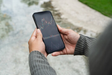 Girl in the park holding a smartphone with Financial stock market graph on the screen. Green area and an artificial lake. Stock Exchange