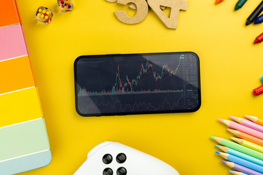 Financial stock market graph on the smartphone screen. Yellow background with school supplies, children's accessories, video game controller. Stock Exchange - Powered by Adobe