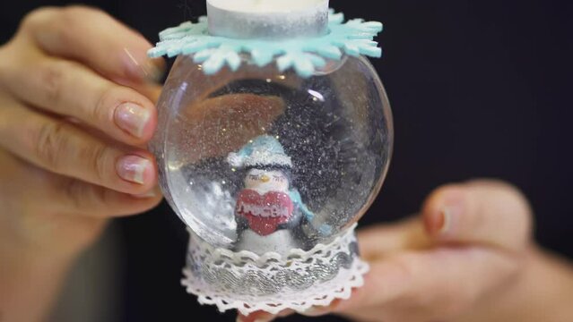 The girl turns over a glass ball with snow in her hands. ART. Inside the snow globe is a penguin with a heart in a scarf