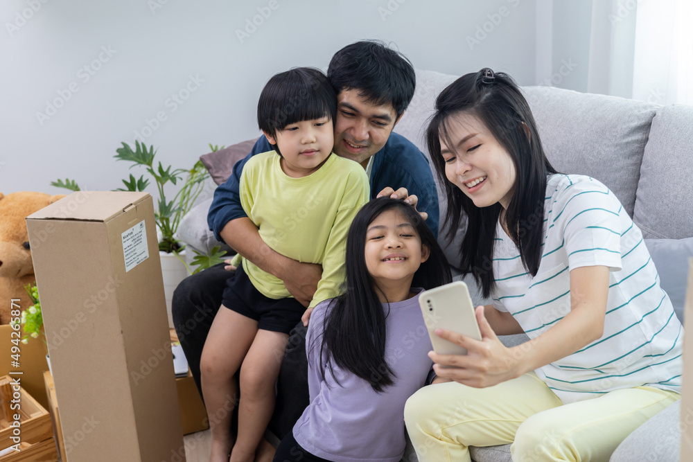 Wall mural Asian family with two little kids sitting on couch at home together, taking selfie, smiling at smartphone camera, mom holding phone. Medium shot. Family photo or communication concept