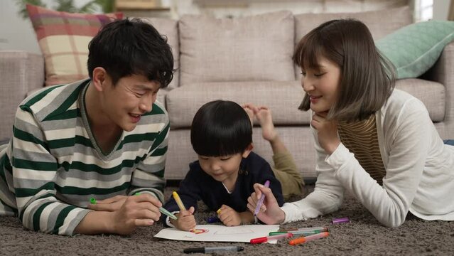 loving asian dad and mom teaching their baby boy how to paint with color pens at home. they lie on belly in the living room at leisure