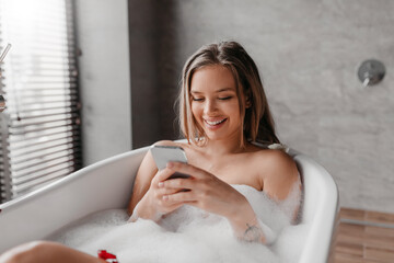 Obraz na płótnie Canvas Happy young lady relaxing in foamy bath, reading beauty blog or watching video online, using smartphone and smiling