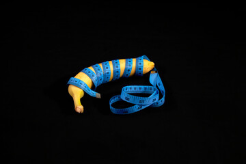 A yellow ripe banana wrapped in blue measure tape, isolated on black. Visual metaphor: checking the...