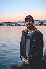 Handsome and happy young man with beard, snapback and jacket standing with sunset over the lake, shipyard and harbor in the back, Valdivia, Chile