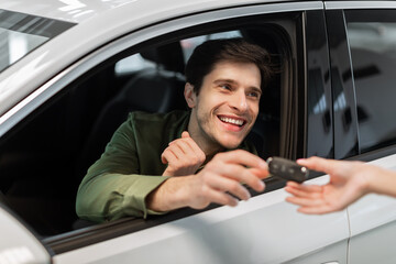 Happy young Caucasian man taking car key from auto dealer, purchasing new vehicle at showroom store