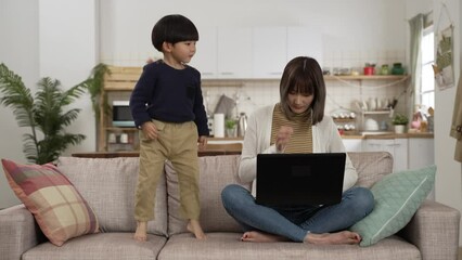 slow motion of stressed Asian mother working from home on the computer while her baby son is playing jump on the sofa. distance working chaos concept - Powered by Adobe