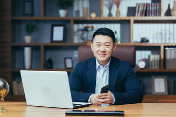 Portrait of a successful Asian in a classic office working on a laptop smiling and looking at the...