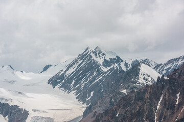 Fototapeta na wymiar Atmospheric landscape with sharp rocks and high snowy mountain top in rainy low clouds at overcast. Dramatic gloomy scenery with large snow mountains and glacier in gray cloudy sky at rainy weather.