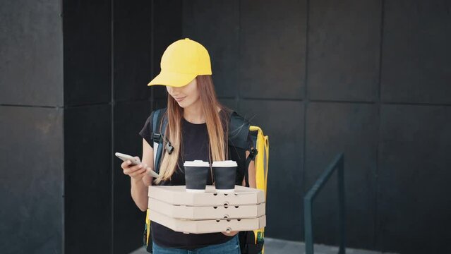 Close up of young woman delivery worker standing at street and holding carton boxes and using smartphone. Delivery Woman stretching out a stack of pizza boxes
