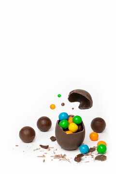 Cracked chocolate easter egg with colorful small round candies and chocolates on white background, copy space. A broken milk chocolate egg with a flying lid. Chocolate Easter concept.