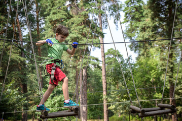 Strong excited young boy playing outdoors in rope park. Caucasian child dressed in casual clothes and sneakers at warm sunny day. Active leisure time with children concept
