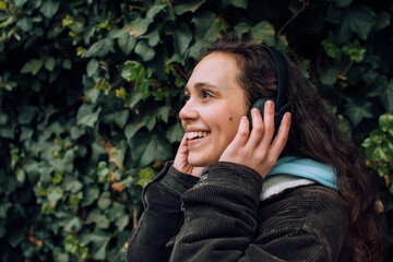 Young girl smiles while listening to music with her headphones in the open air, park, street. Outdoor. Holding his headphones with her hands. Green background, natural, grass. Young model profile.