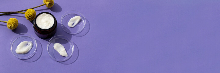 Petri dishes with white cream smears and cream in dark glass jar on violet background. Banner concept