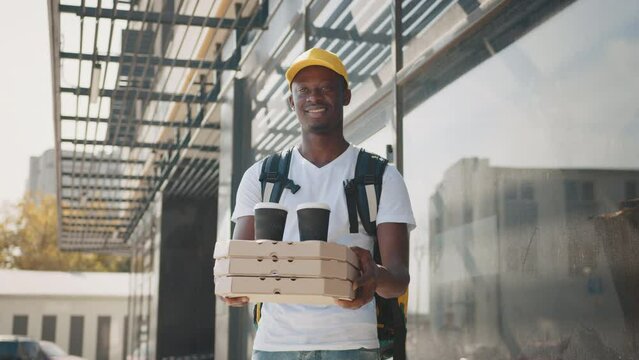 Black Male courier with cardboard boxes delivering coffee cups and pizza to customer while standing outdoors of modern house. African american delivery man carrying pizza boxes and food with coffee.