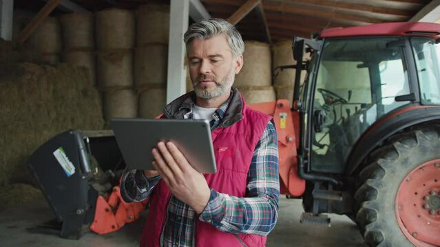Attractive man farmer using digital tablet computer. On background tractor and hay bales. Modern technology in agriculture. Slow motion