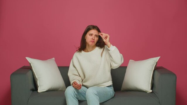 Funny young girl make little size gesture, demonstrate tiny object, shrug shoulders, sitting on couch. Body language