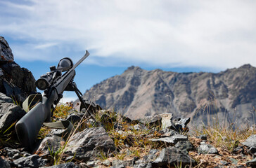 Sniper rifle with optical sight on bipods among rocks in the mountains against the sky.