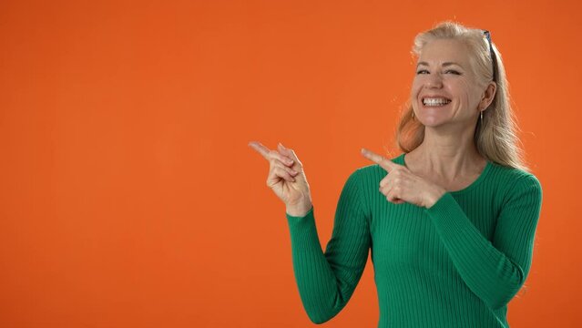 Portrait of pretty mature woman in green shirt smiling showing area to side, pointing to copy space isolated on solid orange background in studio. People emotions lifestyle concept. Mock up