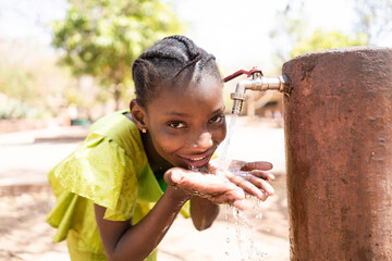 Smiling young African girl at a faucet, letting water flow on her hands, while drinking.