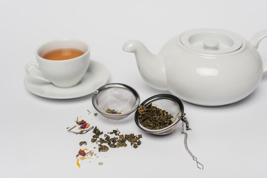 Dry tea in infuser near cup and teapot on white background.