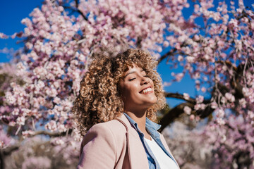 smiling hispanic woman with eyes closed in park enjoying sunny day. Spring flowers background - 494302435
