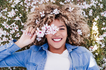 top view of happy hispanic woman with afro hair lying on grass among pink blossom flowers.Springtime
